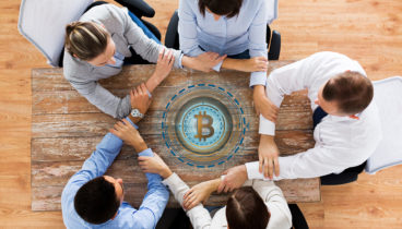 cryptocurrency, cooperation and finance concept concept - business team sitting at office table with bitcoin hologram and holding hands from top